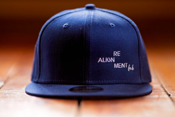 A blue RE.AL NEW ERA® Snapback Hat with the text "RE ALIGN MENT lab" embroidered on the front, displayed on a wooden surface.